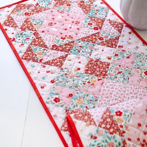 Może przedstawiać: Pikowany bieżnik na stół z kwiatowym wzorem w odcieniach czerwieni, różu i błękitu. Bieżnik ma czerwone obramowanie i jest wykonany z małych, kwadratowych kawałków materiału zszytych razem. Wzór zawiera mieszankę kwiatowych nadruków i jednolitych różowych kwadratów.