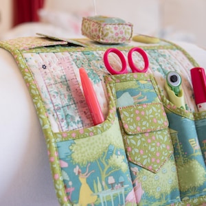 May include: A quilted sewing organizer with multiple pockets, holding pink scissors, a ruler, and a rotary cutter. The organizer features a floral and landscape pattern in shades of green, blue, and pink. A matching pin cushion is also visible.