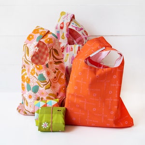 Puede incluir: Tres bolsas de la compra reutilizables hechas de tela. Las bolsas están dobladas y apiladas sobre una superficie blanca. La bolsa de abajo es rosa con un estampado floral. La bolsa del medio es blanca con un estampado colorido de utensilios de cocina. La bolsa de arriba es naranja con un estampado rosa de texto.
