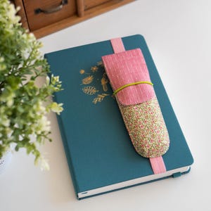 Puede incluir: Una libreta azul verdosa con un diseño floral dorado en la portada. Un porta lápices de tela rosa y verde está unido a la libreta con una banda elástica verde.