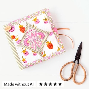 May include: A square fabric needle case featuring a white background with a strawberry print, a central diamond-shaped floral patch, a pink button closure with leather ties, and a small wooden tag labeled handmade, accompanied by metal scissors.
