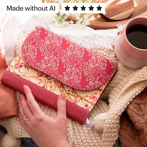 May include: A fabric book cover featuring a red flap with a white speckled pattern and a cream-colored body decorated with a floral branch print. The cover includes a solid red ribbed fabric band across the bottom and a small light purple ribbon bookmark.