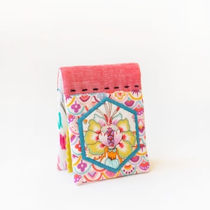 Puede incluir: Una pequeña bolsa de tela rosa y blanca con un diseño floral hexagonal en el centro. La bolsa tiene una solapa superior roja y está decorada con un ribete de cuentas turquesa.