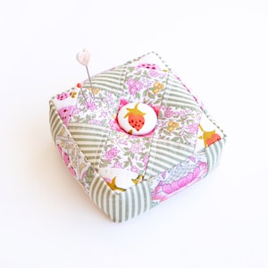 May include: A green and white patchwork pincushion with a pink flower and a strawberry button in the centre. The pincushion has a heart-shaped pin stuck in it.