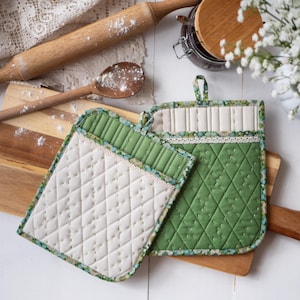 Może przedstawiać: Dwa pikowane uchwyty do garnków, jeden zielony, a drugi biały z zielonym wykończeniem. Oba mają kwiatowy wzór z tyłu.