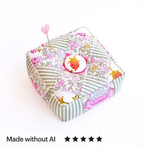 以下が含まれることがあります： ピンク、緑、白の花柄パッチワークデザインと緑のストライプが特徴の正方形の布製ピンクッション。中央には赤いイチゴのイラストが描かれた白いボタンがあり、ピンクのハート型の頭が付いたピンが刺さっています。
