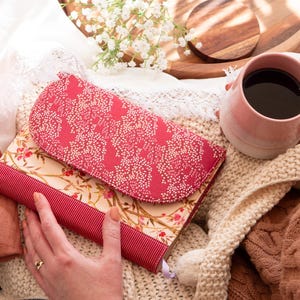 May include: A pink and white floral patterned journal with a red ribbon closure. The journal is sitting on a white knitted blanket with a pink mug of coffee.
