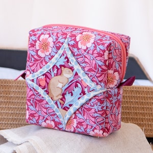 Pode incluir: Uma bolsa de cosméticos de tecido rosa e azul com fecho de correr. A bolsa apresenta um design acolchoado com um esquilo a dormir no centro.