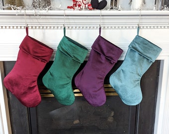 Op de afbeelding: Vier fluwelen kerstsokken hangen aan een witte schoorsteenmantel. De sokken zijn bordeauxrood, teal, groen en paars.
