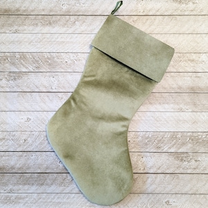 May include: A sage green Christmas stocking with a folded cuff and a hanging loop. The stocking is made of a soft, velvety material and is displayed against a light wood-grain background. The stocking is approximately 18 inches long.