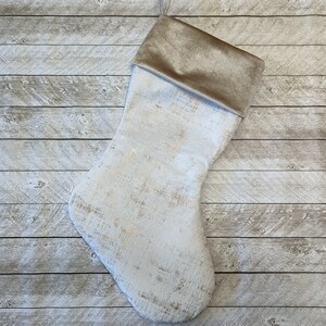 May include: A Christmas stocking with a white and gold distressed pattern. The cuff is a solid tan colour. The stocking is made of soft fabric and is designed for holiday decor.