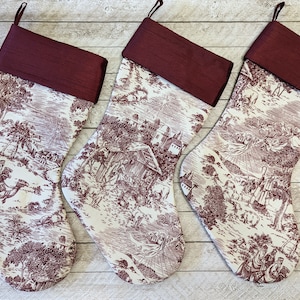 Puede incluir: Tres calcetines de Navidad rojos y blancos con un patrón de toile. Los calcetines tienen un puño de satén burdeos.