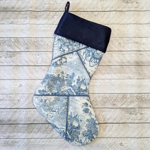 May include: A blue and white Christmas stocking with a black velvet cuff. The stocking features a toile pattern with a repeating design of houses, trees, and flowers.