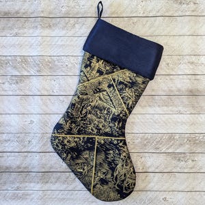 Peut inclure: Une chaussette de Noël noire et dorée avec un motif décoratif. La chaussette a un revers noir et un motif doré qui ressemble à une scène d'un village médiéval.