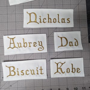 Puede incluir: Cinco etiquetas de tela blanca con texto bordado en oro. Las etiquetas dicen "Nicholas", "Aubrey", "Dad", "Biscuit" y "Kobe".