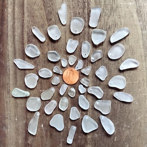 Op de afbeelding: Een verzameling van 30 witte stukjes zeeglas, gerangschikt in een cirkel rond een cent. Het zeeglas is glad en doorschijnend, met een verscheidenheid aan vormen en maten.