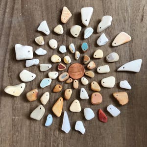 Puede incluir: Una colección de 30 piezas de vidrio de mar liso, blanco y marrón dispuestas en círculo sobre una superficie de madera. Las piezas de vidrio de mar son de diversas formas y tamaños, algunas con un agujero perforado. Un centavo de cobre se encuentra en el centro del círculo.