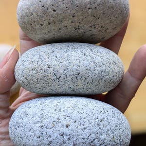 May include: Three smooth, gray river stones stacked vertically, held in a hand. The stones have a speckled texture and varying sizes. The background is blurred, suggesting an outdoor setting.