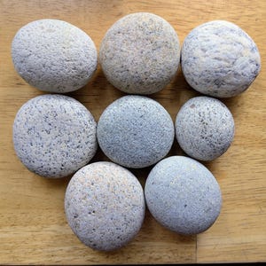 May include: An overhead shot of nine smooth, rounded stones in varying shades of gray and tan. The stones are arranged on a light brown wooden surface, showcasing their natural textures and colors.