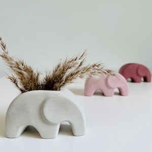 May include: Three ceramic elephant planters in different shades of pink and gray. The gray planter is the largest and has a small bouquet of dried brown grass inside.