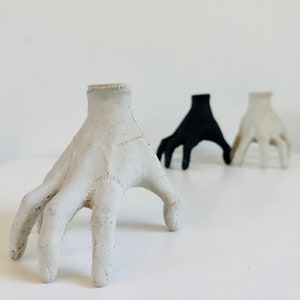 May include: Three concrete hand sculptures, one white, one black, and one gray, with outstretched fingers. The sculptures are arranged in a row on a white surface.