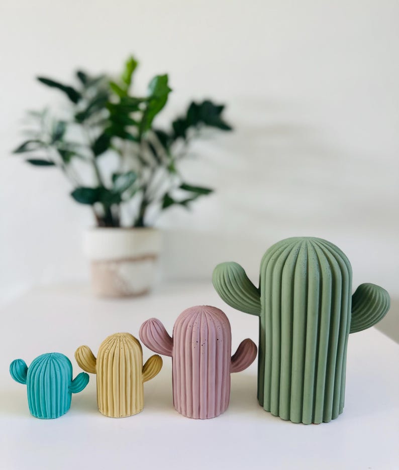 May include: Four different sized concrete cactus figurines in various colors. The largest cactus is green, the next is pink, then yellow, and the smallest is blue.
