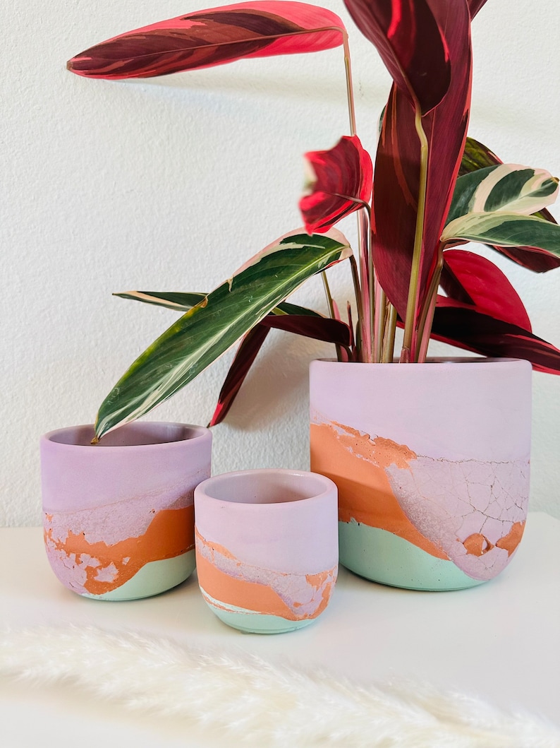 May include: Three different sized, hand-painted ceramic planters in shades of purple, orange, and green. The planters are displayed on a white surface with a fluffy white rug in the foreground.