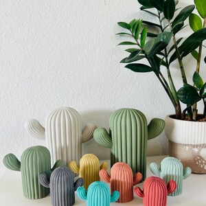 May include: A set of nine colorful ceramic cactus figurines in various sizes. The cacti are painted in shades of green, gray, teal, yellow, red, and pink. The figurines are arranged on a white surface.