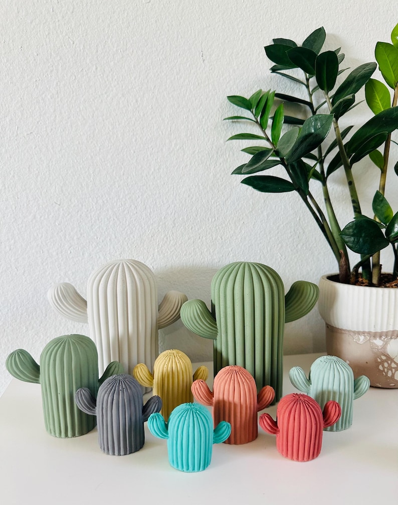 May include: A set of nine colorful ceramic cactus figurines in various sizes. The cacti are painted in shades of green, gray, teal, yellow, red, and pink. The figurines are arranged on a white surface.
