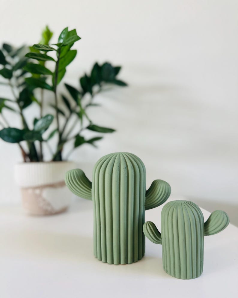 May include: Two green ceramic cactus sculptures with a ribbed texture. The larger cactus is taller and has two arms, while the smaller cactus has one arm.