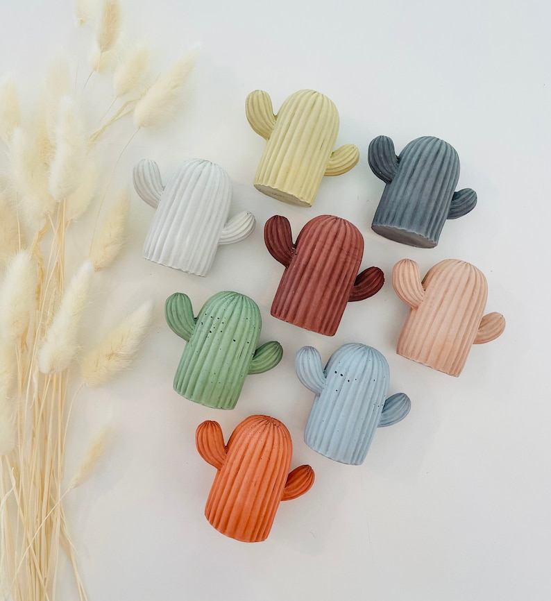 May include: Nine small concrete cactus figurines in various colors, including yellow, white, gray, red, green, light blue, and orange. The cacti are all in the same style, with a rounded body and two arms.
