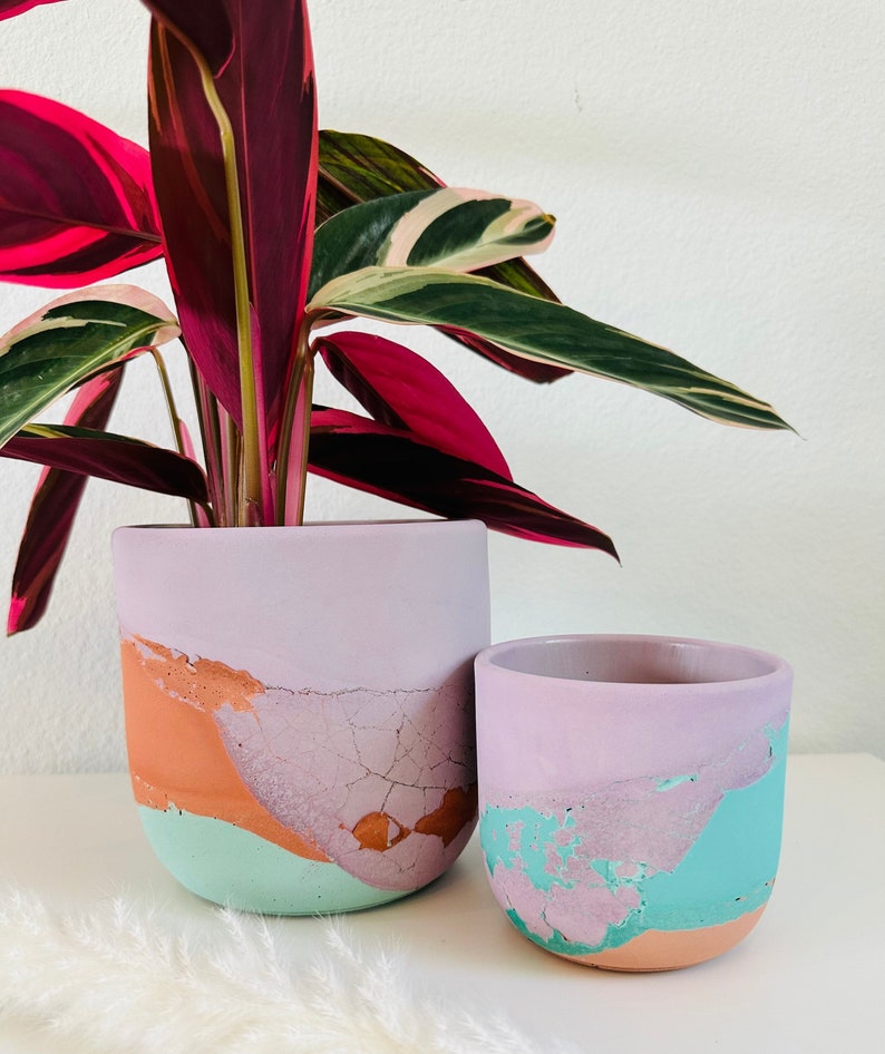 May include: Two colorful concrete planters with a distressed, crackled finish. The larger planter is light purple with orange, green, and pink accents. The smaller planter is light purple with green and orange accents.