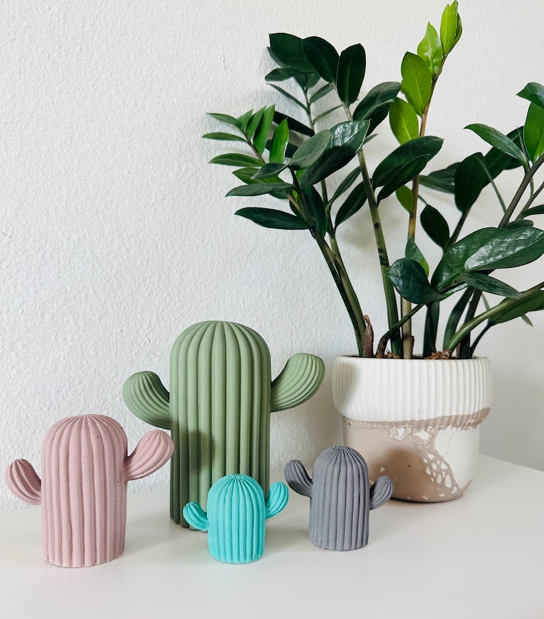 May include: Four small cactus figurines in different colors, including green, pink, blue, and gray, are arranged on a white surface. A larger green cactus figurine is behind the smaller ones. A potted plant with green leaves is in the background.