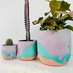 May include: Three different sized concrete planters with a marbled effect in shades of pink, teal, and brown. The planters are filled with plants, including a cactus and a leafy green plant.
