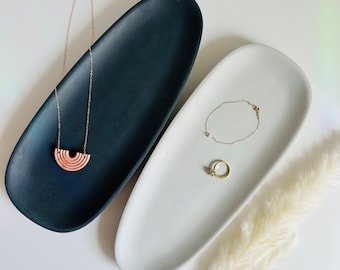 May include: Two oval ceramic dishes, one black and one white, with jewelry on top. The black dish has a pink and gold necklace with a rainbow design. The white dish has a gold bracelet and a gold ring.