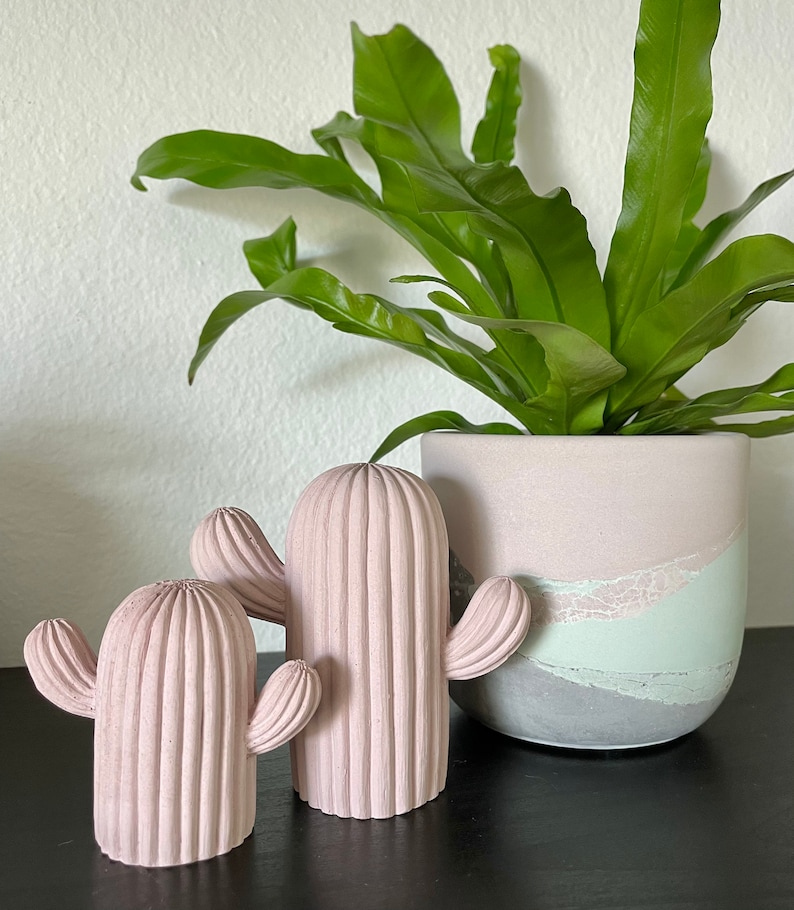 May include: Two pink concrete cactus sculptures with a green fern plant in a pink and gray planter.