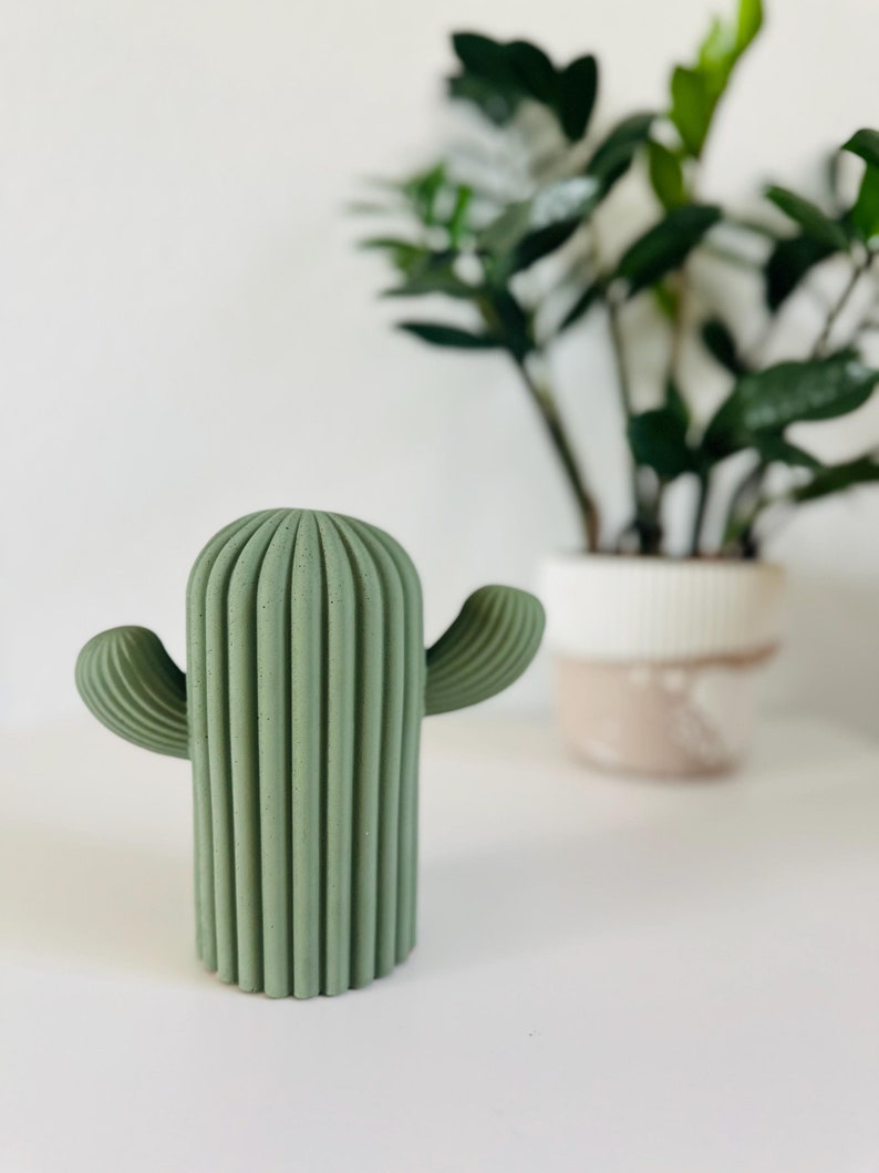 May include: A green concrete cactus sculpture with a ribbed texture and two arms. The cactus is sitting on a white surface.