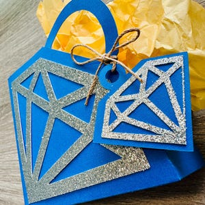 May include: A blue gift bag featuring a diamond-shaped cutout design, embellished with silver glitter. A matching tag is attached with twine. The bag is filled with yellow tissue paper, creating a festive presentation.