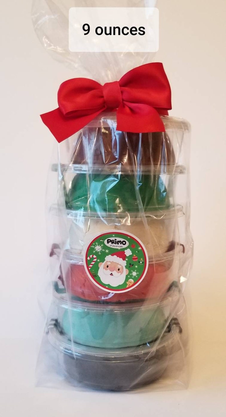 Op de afbeelding: Een stapel van vijf bakjes met klei, verpakt in doorzichtig plastic met een rode strik. De bakjes zijn in een piramidevorm gestapeld. De klei is in de kleuren bruin, groen, wit, rood en groen. De bakjes hebben een etiket met een cartoon kerstman. Het etiket zegt "Primo Playdough". De tekst "9 ounces" staat op een wit etiket bovenaan de afbeelding.