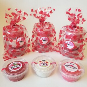 May include: Three clear plastic bags with red hearts printed on them, each containing three small plastic containers of pink and white slime. The containers are labeled with the words "Strawberry Berry", "Love Potion", and "Cupcake".