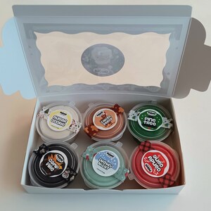 Op de afbeelding: Een set van zes kleine potjes met kleurrijke klei met feestelijke, kerstthema-etiketten. De etiketten omvatten "Sugar Cookie", "Gingerbread", "Pine Tree", "Camp Fire", "Snowy Night" en "Candy Cane".