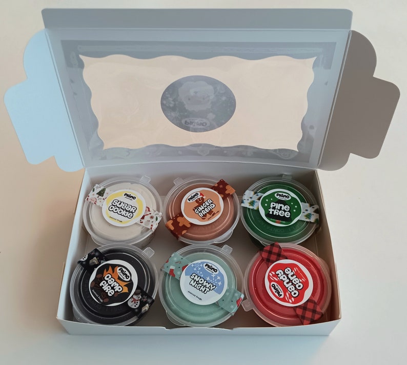 Op de afbeelding: Een set van zes kleine potjes met kleurrijke klei met feestelijke, kerstthema-etiketten. De etiketten omvatten "Sugar Cookie", "Gingerbread", "Pine Tree", "Camp Fire", "Snowy Night" en "Candy Cane".