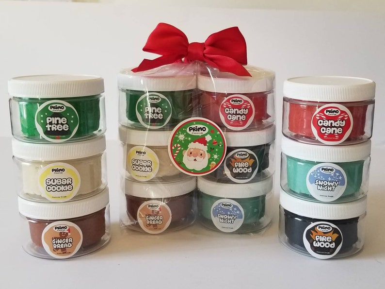 Op de afbeelding: Een set van 10 kleine potjes met klei, elk met een andere kerstgeur en kleur. De potjes zijn gerangschikt in een cadeauzak met een rode strik. De geuren zijn onder andere dennenboom, suikerkoekje, snoepstok, houtvuur en sneeuwwinter.
