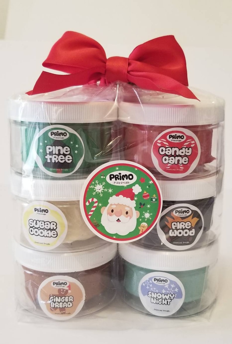 Op de afbeelding: Een set van zes kerstthema playdough-containers in een doorzichtige plastic zak met een rode strik. De containers zijn gestapeld in twee rijen van drie. De playdough-smaken zijn Pine Tree, Candy Cane, Sugar Cookie, Fire Wood, Gingerbread en Snowy Night.