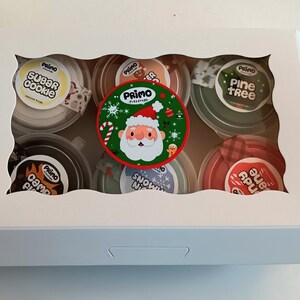 Op de afbeelding: Een witte doos met een transparant venster dat zes kleine containers met slijm laat zien. De containers zijn versierd met kerstthema-ontwerpen en labels met de tekst "Sugar Cookie", "Camp Fire", "Snow Night", "Pine Tree", "Candy Cane" en "Gingerbread".