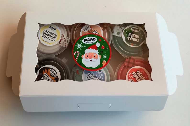 Op de afbeelding: Een witte doos met een transparant venster dat zes kleine containers met slijm laat zien. De containers zijn versierd met kerstthema-ontwerpen en labels met de tekst "Sugar Cookie", "Camp Fire", "Snow Night", "Pine Tree", "Candy Cane" en "Gingerbread".