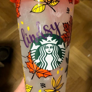 May include: A clear reusable cup with a red lid and a teal straw. The cup features a fall-themed design with orange, yellow, and black leaves. The Starbucks logo is in the centre of the cup, and the name "Lindsey" is written in purple on the side.