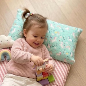 May include: A child plays with a wooden unicorn stacking toy. The child is wearing a pink sweater and white pants, lying on a striped pink and white mat. A pillow with a unicorn pattern is behind the child. A rainbow and cloud toy are also visible.