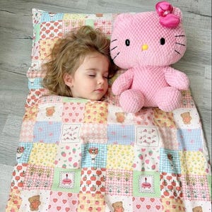 May include: A patchwork quilt featuring a grid of pastel-colored squares with patterns of teddy bears, hearts, strawberries, flowers, and cakes. A large, textured pink plush cat toy with a bright pink bow sits on the quilt next to a sleeping child.