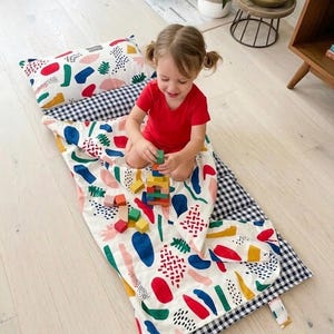 Könnte beinhalten: Ein Kinderschlafsack-Set mit Kissen, mit einem farbenfrohen abstrakten Muster auf weißem Hintergrund. Der Schlafsack hat einen schwarz-weiß karierten Rand. Das Kind trägt ein rotes T-Shirt und spielt mit Holzblöcken.